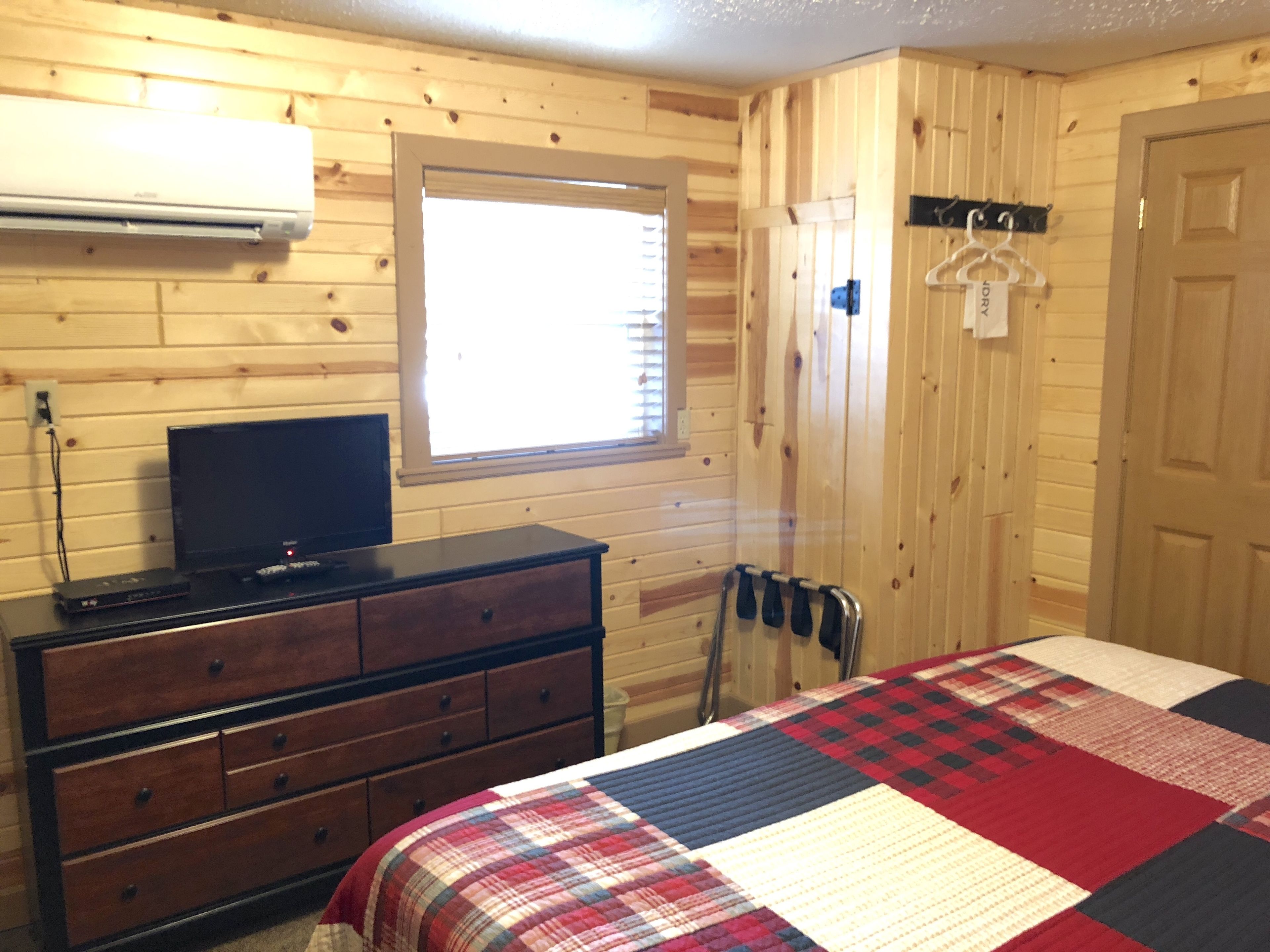 Una habitación de cabaña de madera con cama, cómoda, televisor y ventana con persianas.