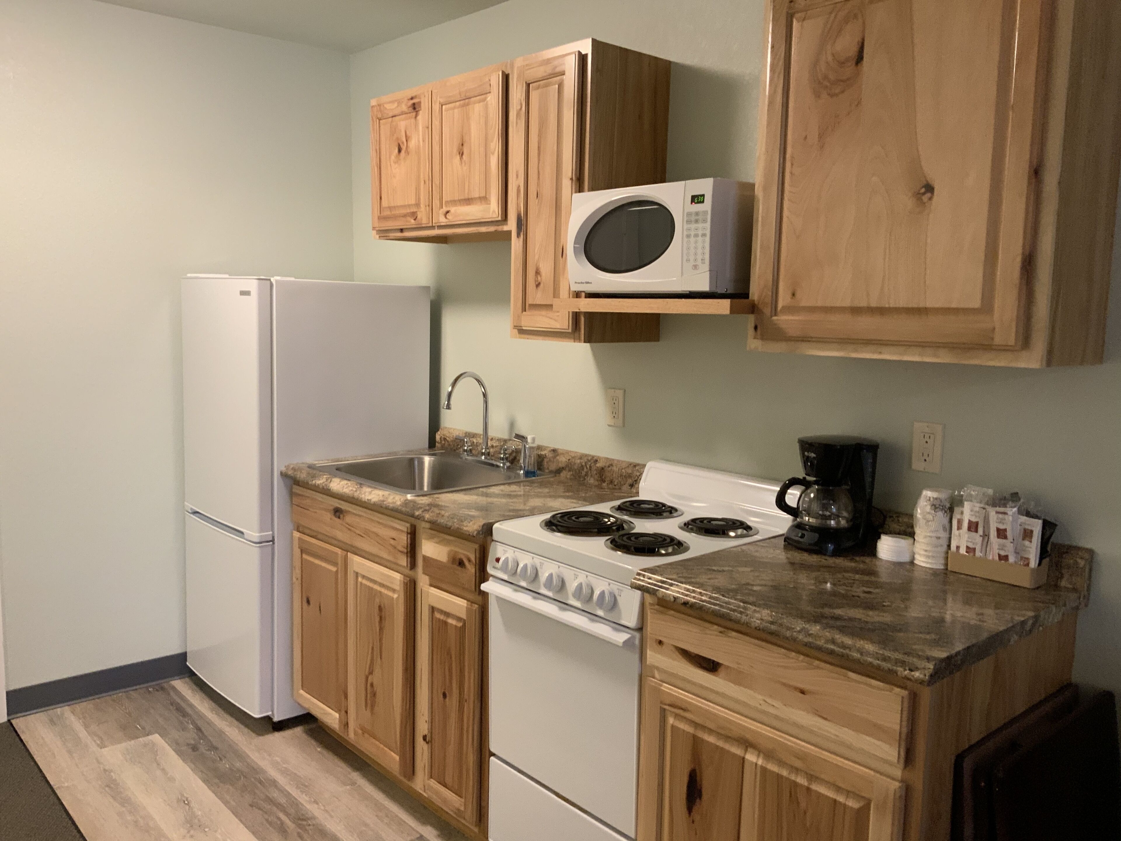 A compact kitchen with wooden cabinets, a white refrigerator, a microwave, a stove, and a coffee maker.