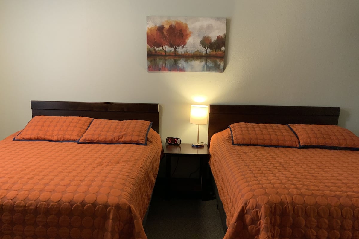 Une chambre d’hôtel avec deux lits, chacun recouvert d’une courtepointe matelassée orange et agrémenté d’oreillers assortis. Une table de chevet avec une lampe et une horloge numérique se trouve entre les deux lits. Au-dessus de la table de chevet, on peut voir un tableau encadré représentant des arbres et une étendue d’eau.