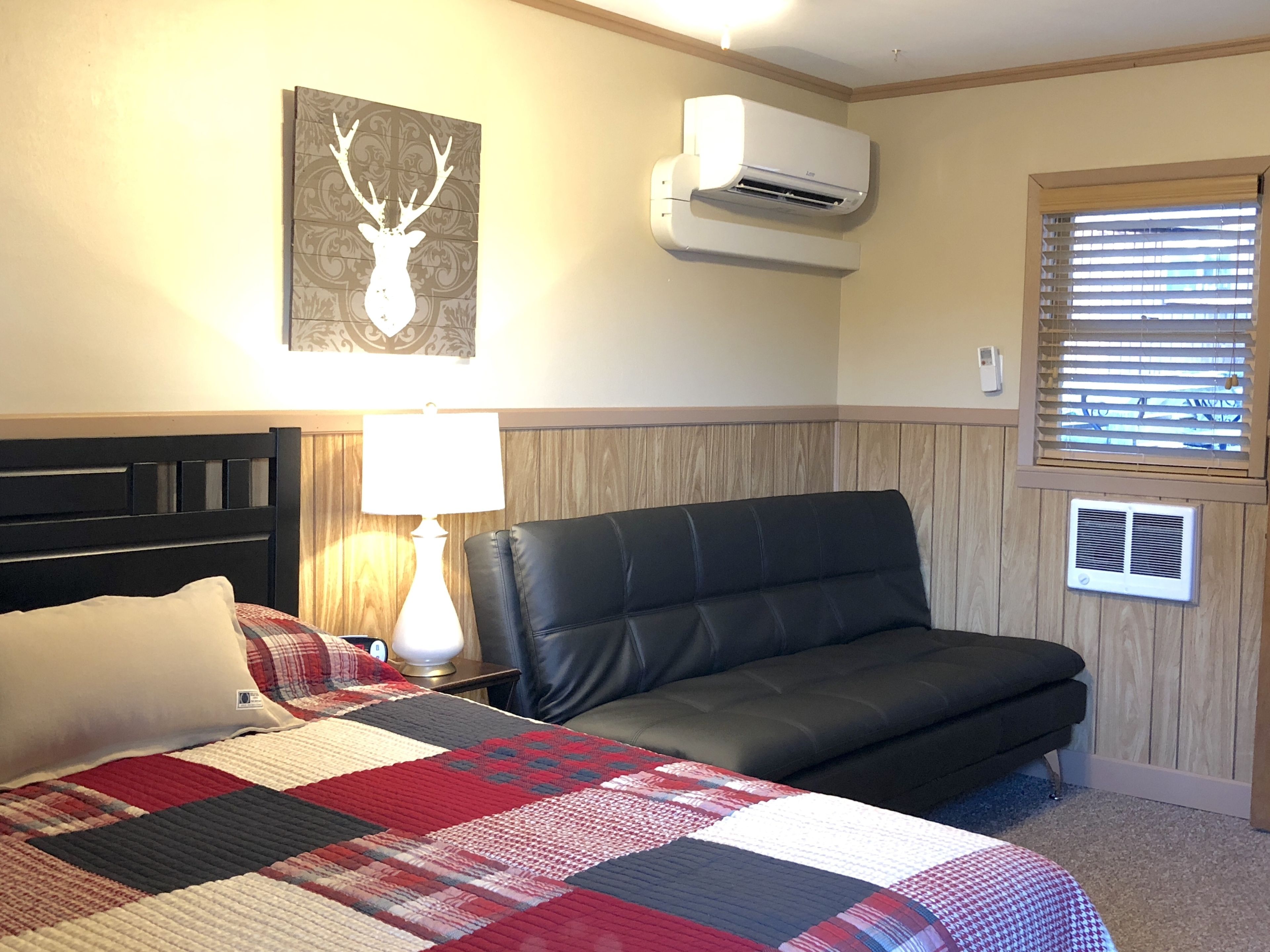 Een slaapkamer met een bed, een zwarte leren bank, een sprei, een wandairconditioner en een raam met jaloezieën.
