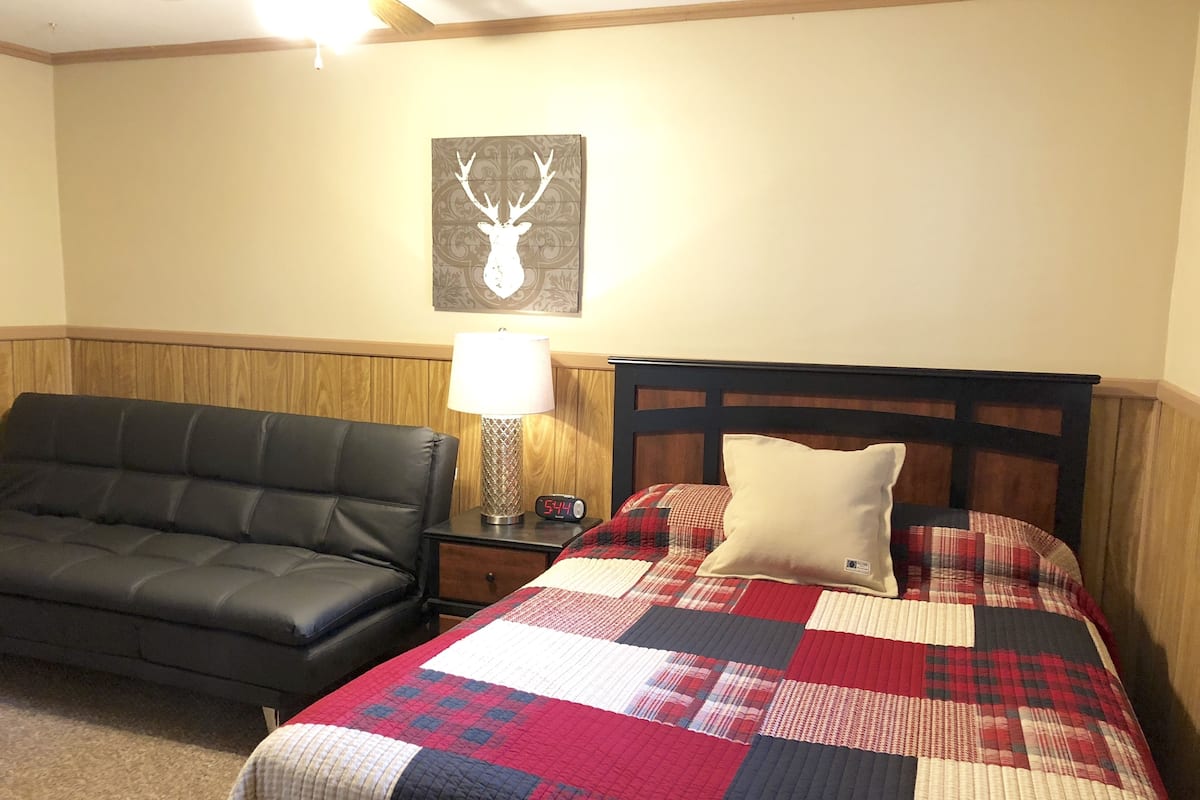 Une chambre avec un canapé en cuir noir, un lit avec une tête de lit en bois, une couvre-lit matelassée, une table de chevet avec une lampe et une décoration murale représentant une tête de cerf.