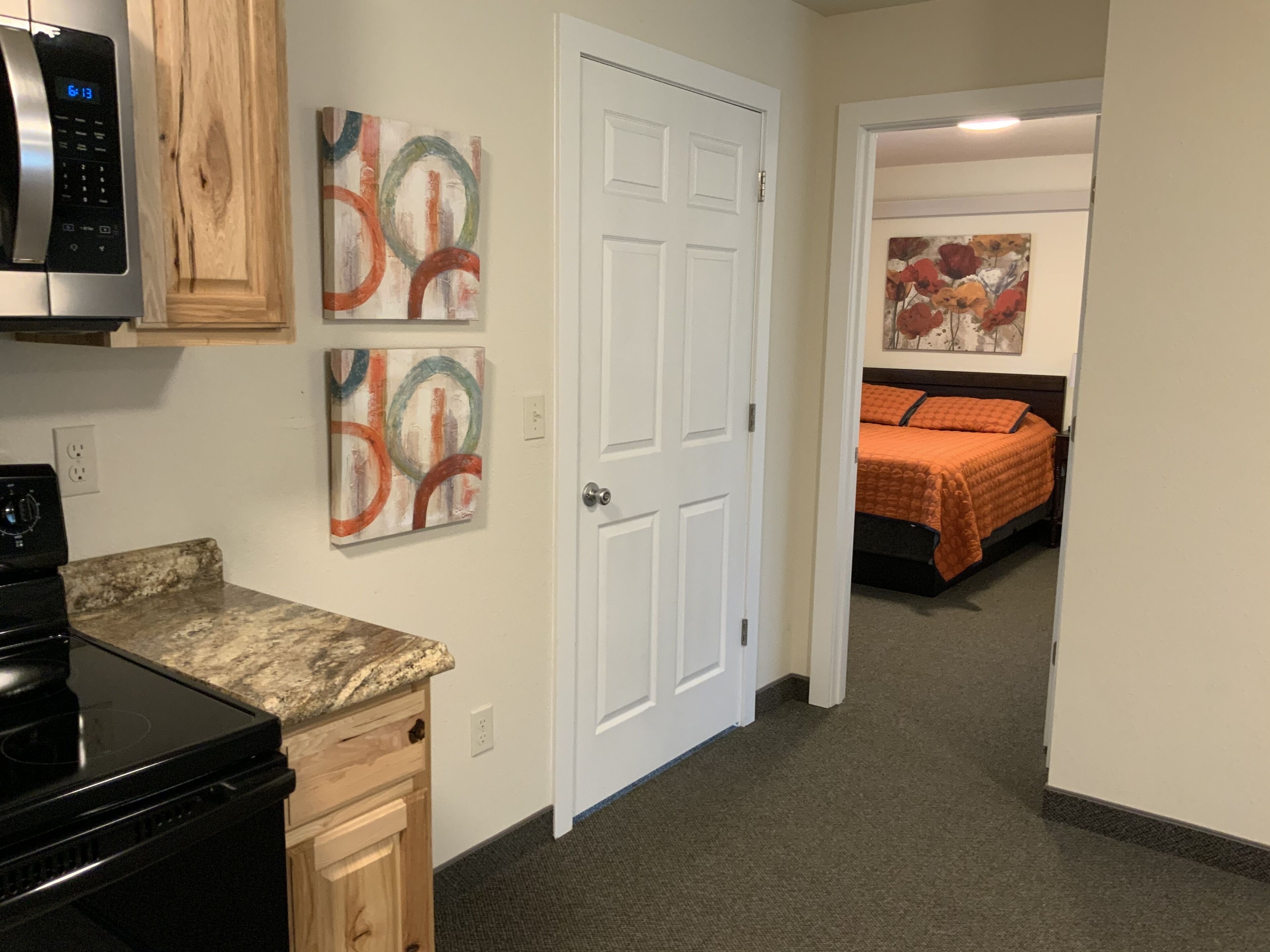 A compact kitchen with a microwave, stove, and wooden cabinets. There are abstract paintings on the wall and a door leading to a bedroom with a bed and a painting.