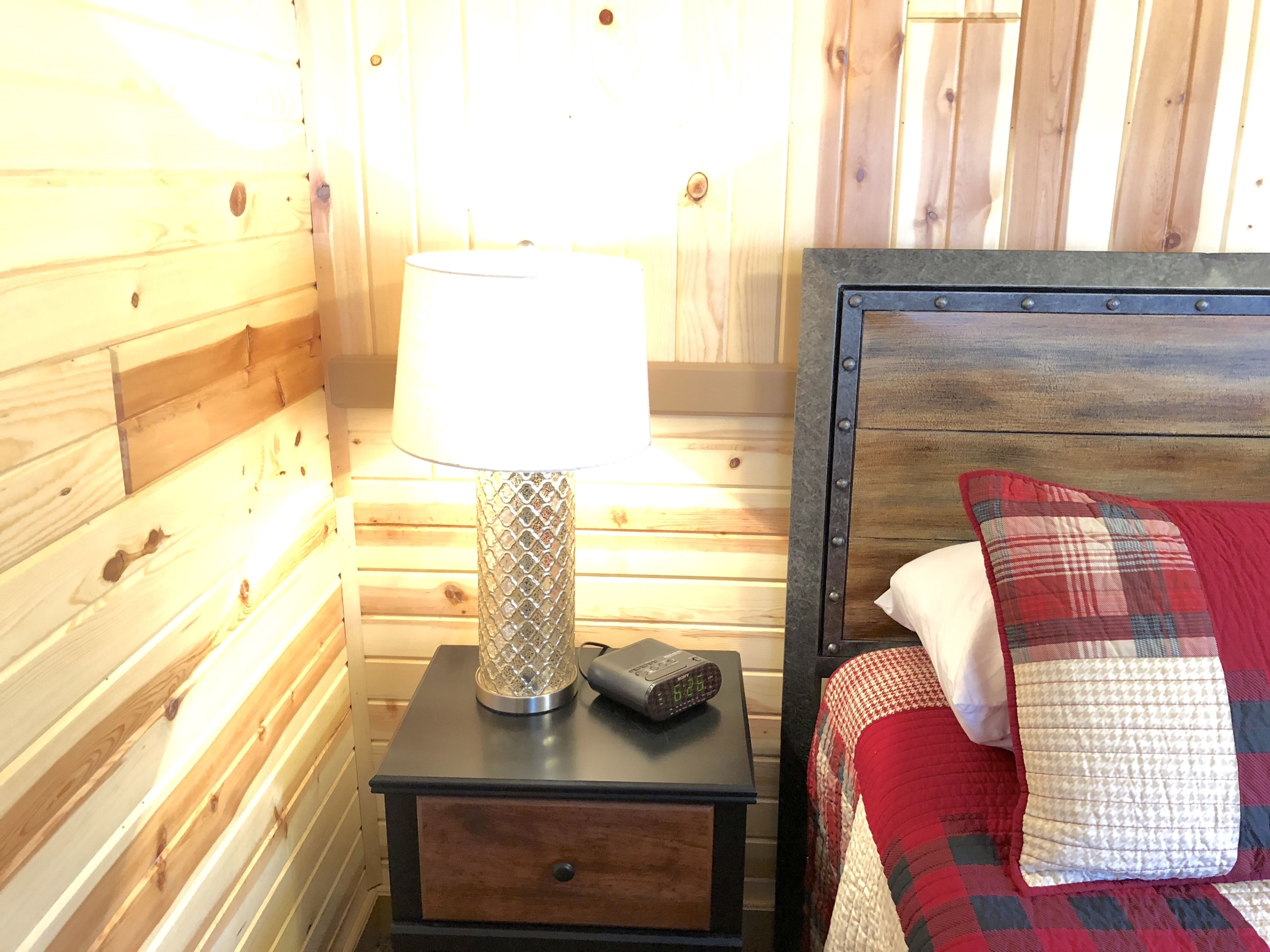 Habitación de cabaña de madera con mesita de noche, lámpara, reloj y cama con manta y almohadas a cuadros.