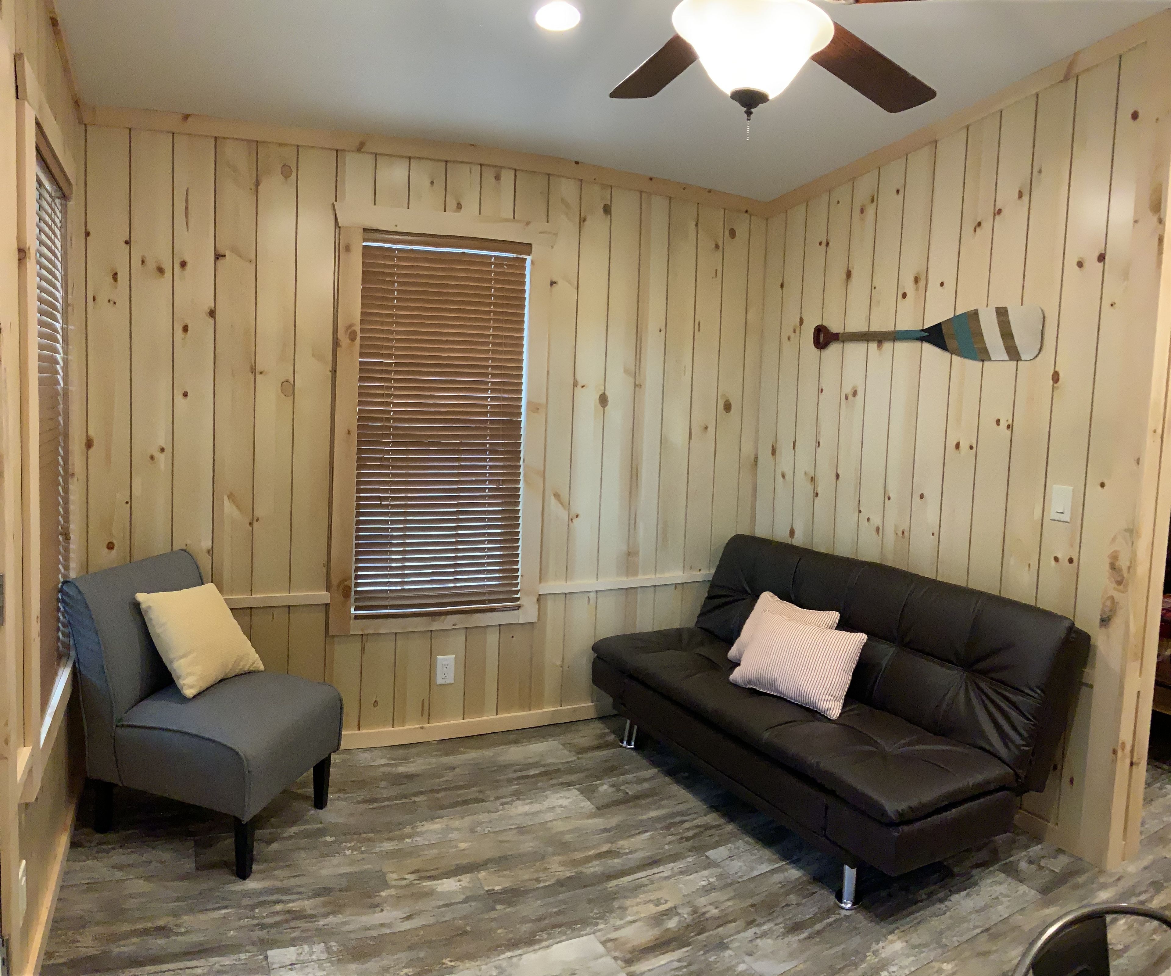 Une pièce aux murs recouverts de lambris en bois, avec un ventilateur de plafond, une fenêtre équipée de stores, un fauteuil gris agrémenté d’un coussin et un canapé marron avec un coussin.