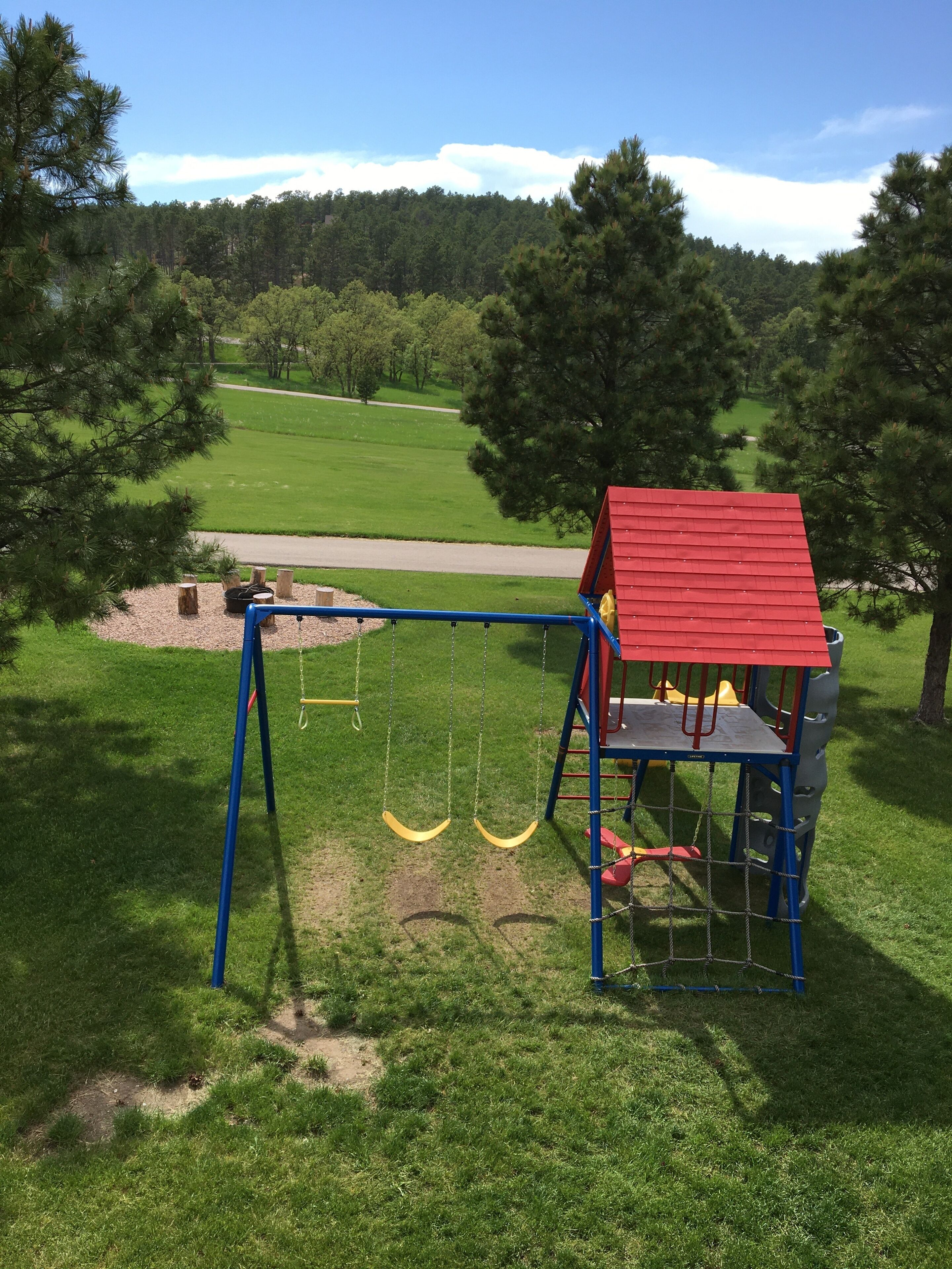 A playground with swings and a climbing structure, surrounded by grass and trees.