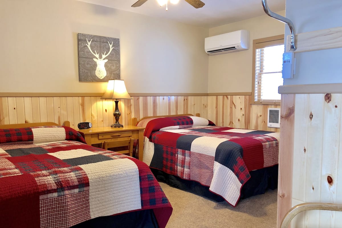 Une chambre avec deux lits, une tête de lit en bois, un climatiseur mural, une lampe et une décoration murale représentant une tête de cerf.