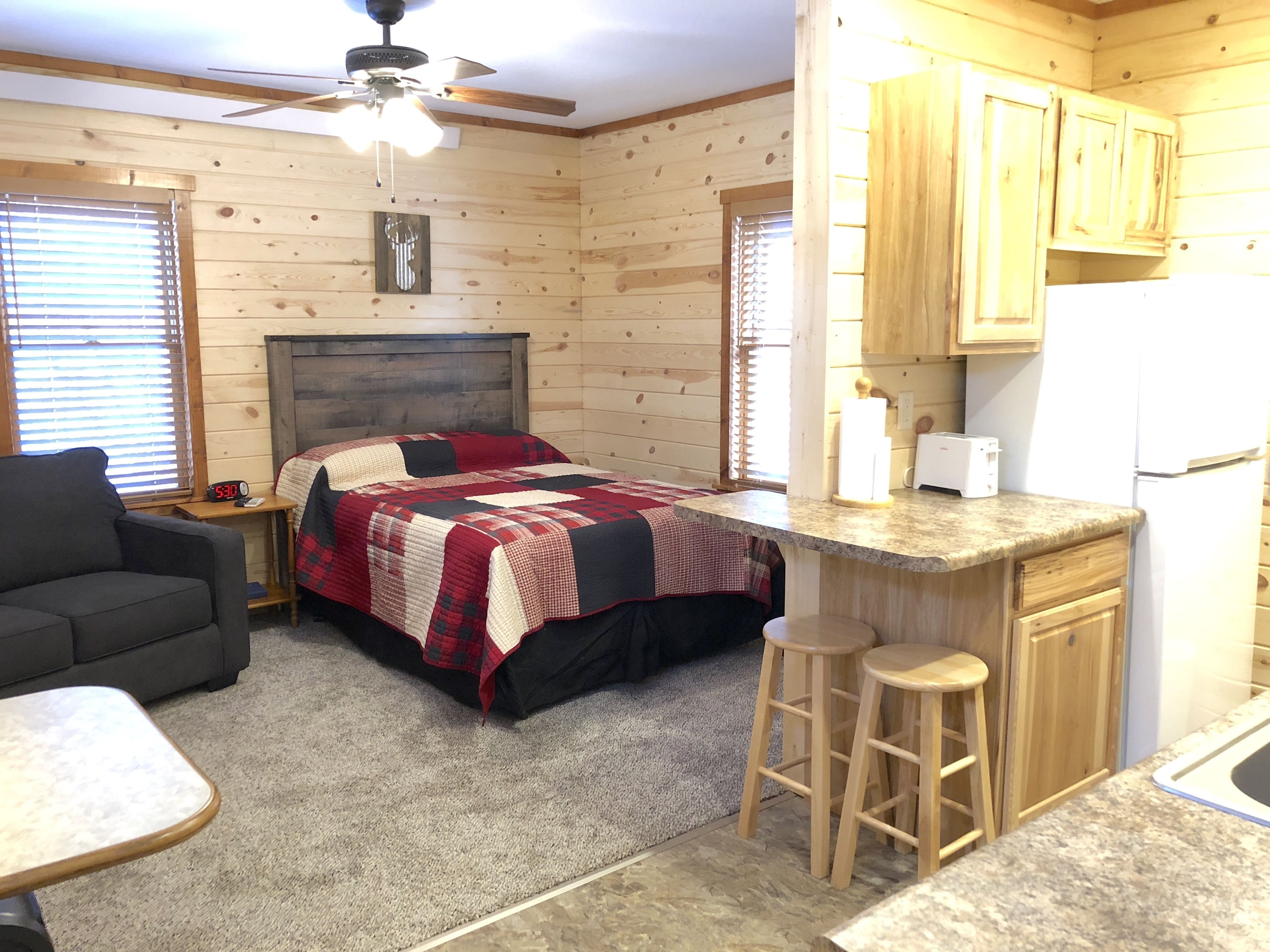 Ein gemütliches Blockhaus mit Bett, einer kleinen Küchenzeile, einem Essbereich und einem Deckenventilator.
