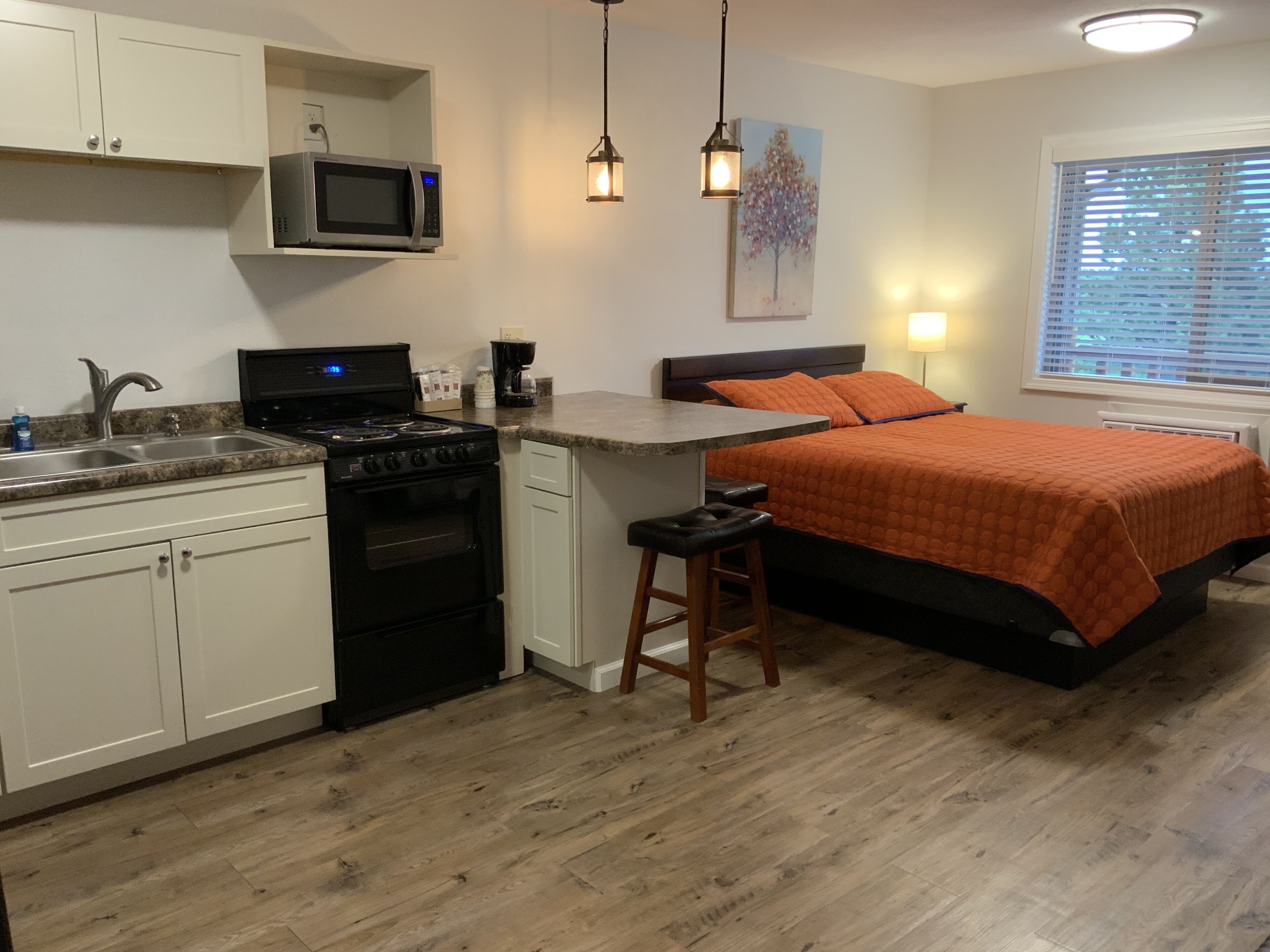 Una cocina compacta con microondas, estufa y lavamanos, un taburete pequeño y una cama con colcha naranja.