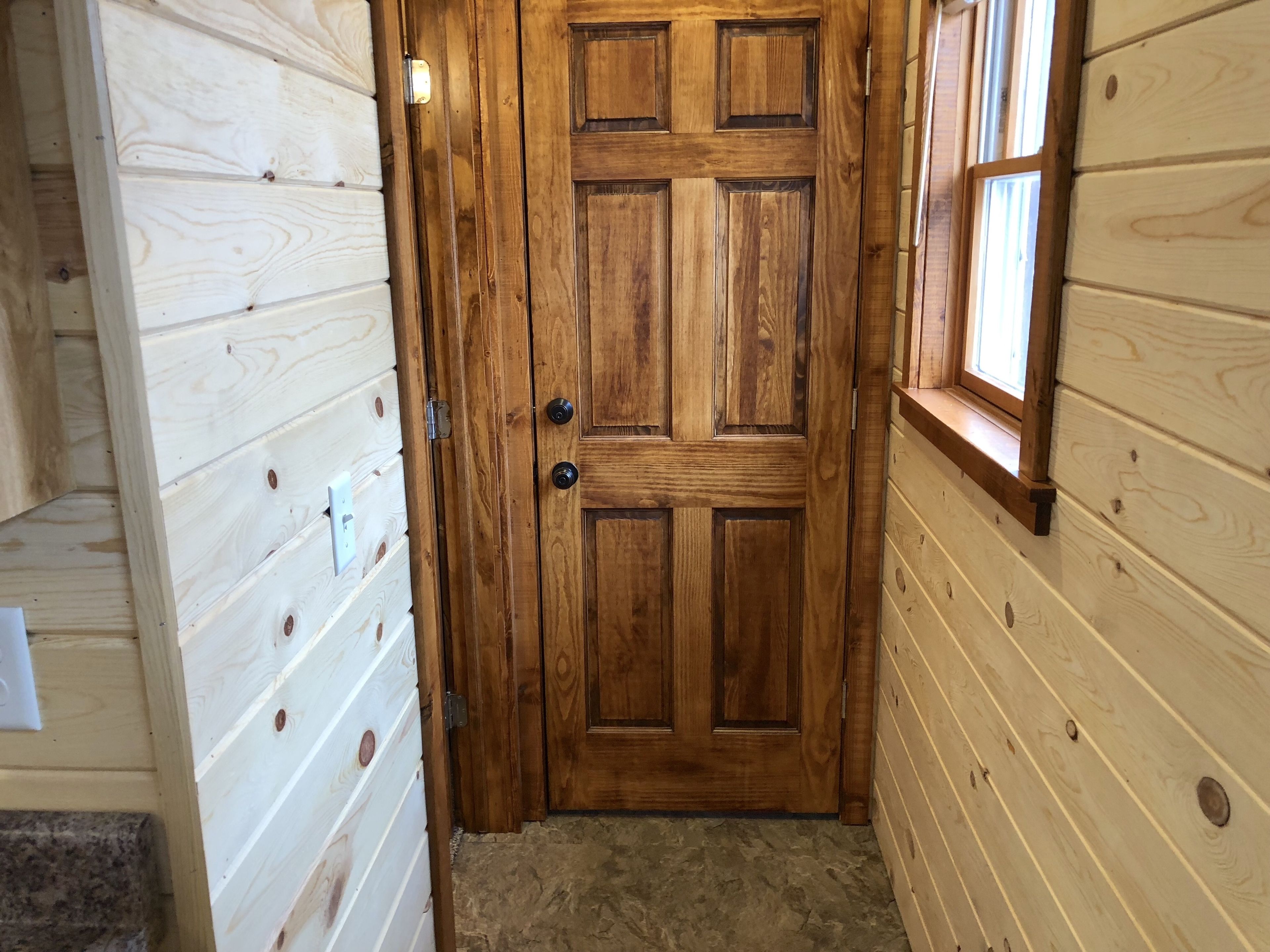 Una porta in legno con una finestra a destra e un interruttore della luce a sinistra, su un pavimento rivestito di moquette.