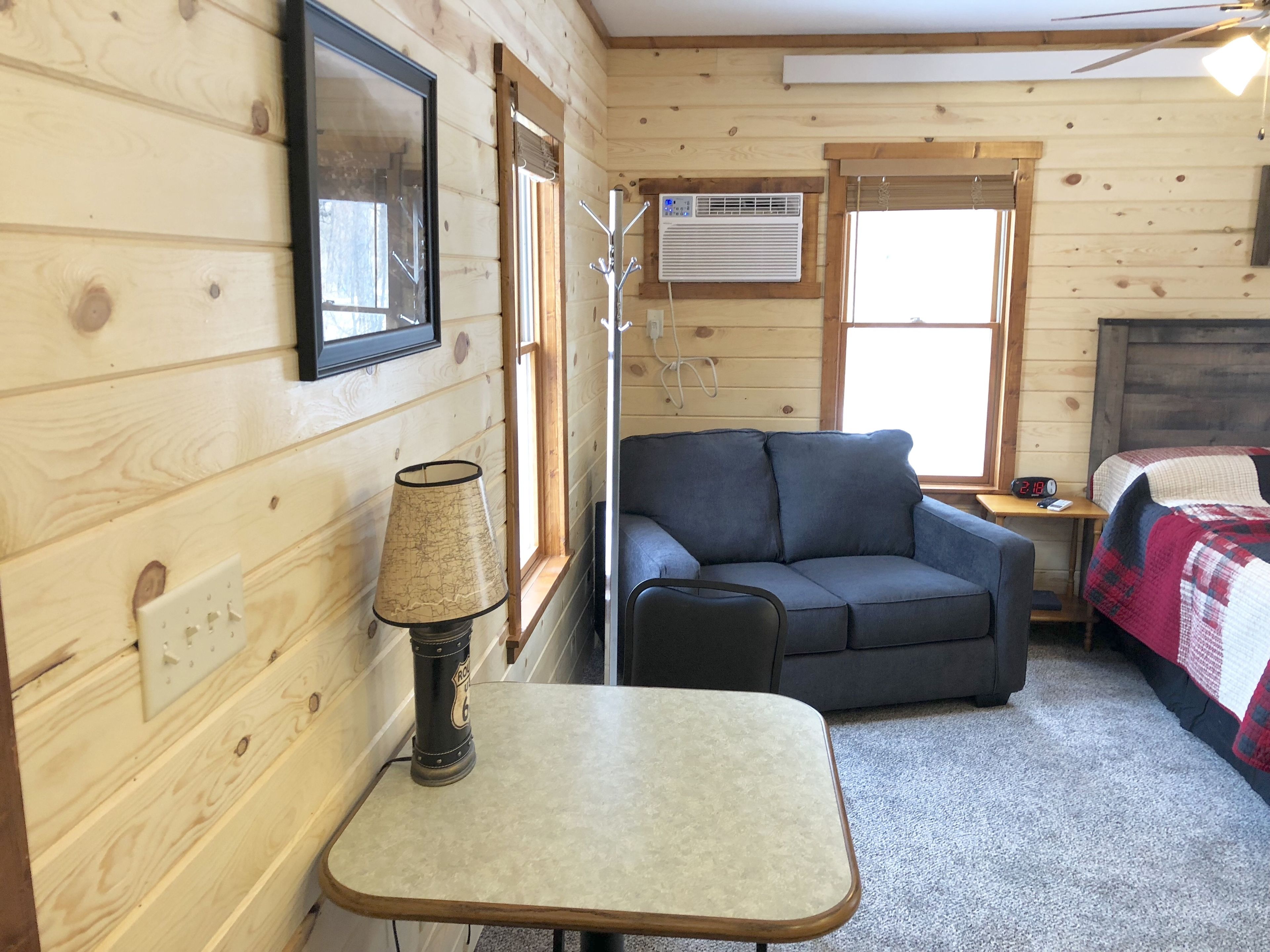 A cozy room with a sofa, a small table, a lamp, a bed with a quilt, and an air conditioning unit.