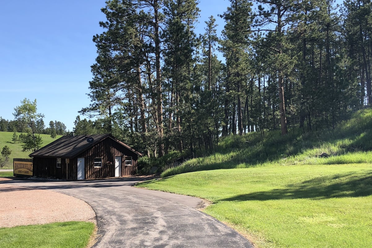 Un chalet rustique en bois, avec une allée de gravier menant à l’entrée, entouré d’un paysage verdoyant et luxuriant et de grands pins.