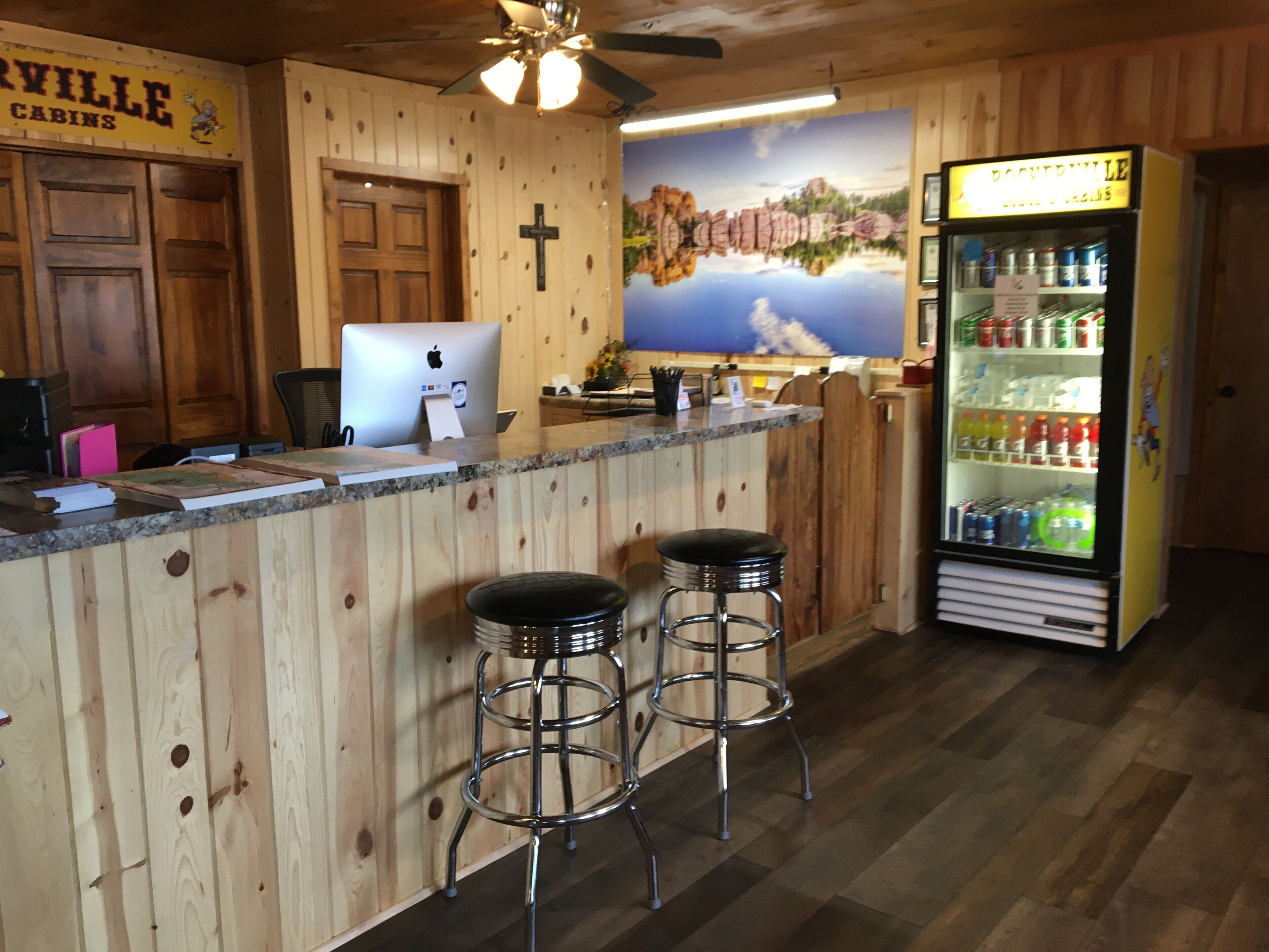 Zona de bar estilo cabaña de madera, con una computadora, dos taburetes y un refrigerador lleno de bebidas.