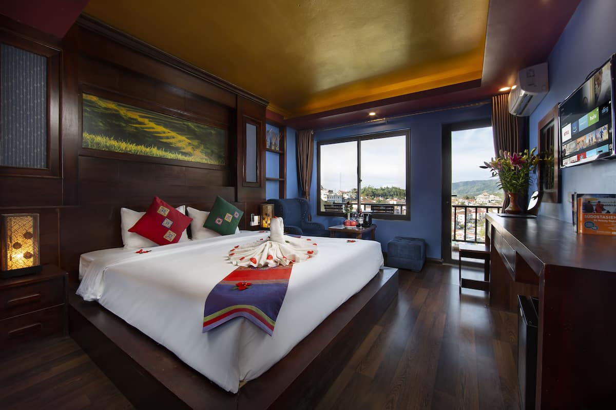 Habitación de hotel con una cama grande, vistas de la ciudad, un balcón y un televisor en la pared.