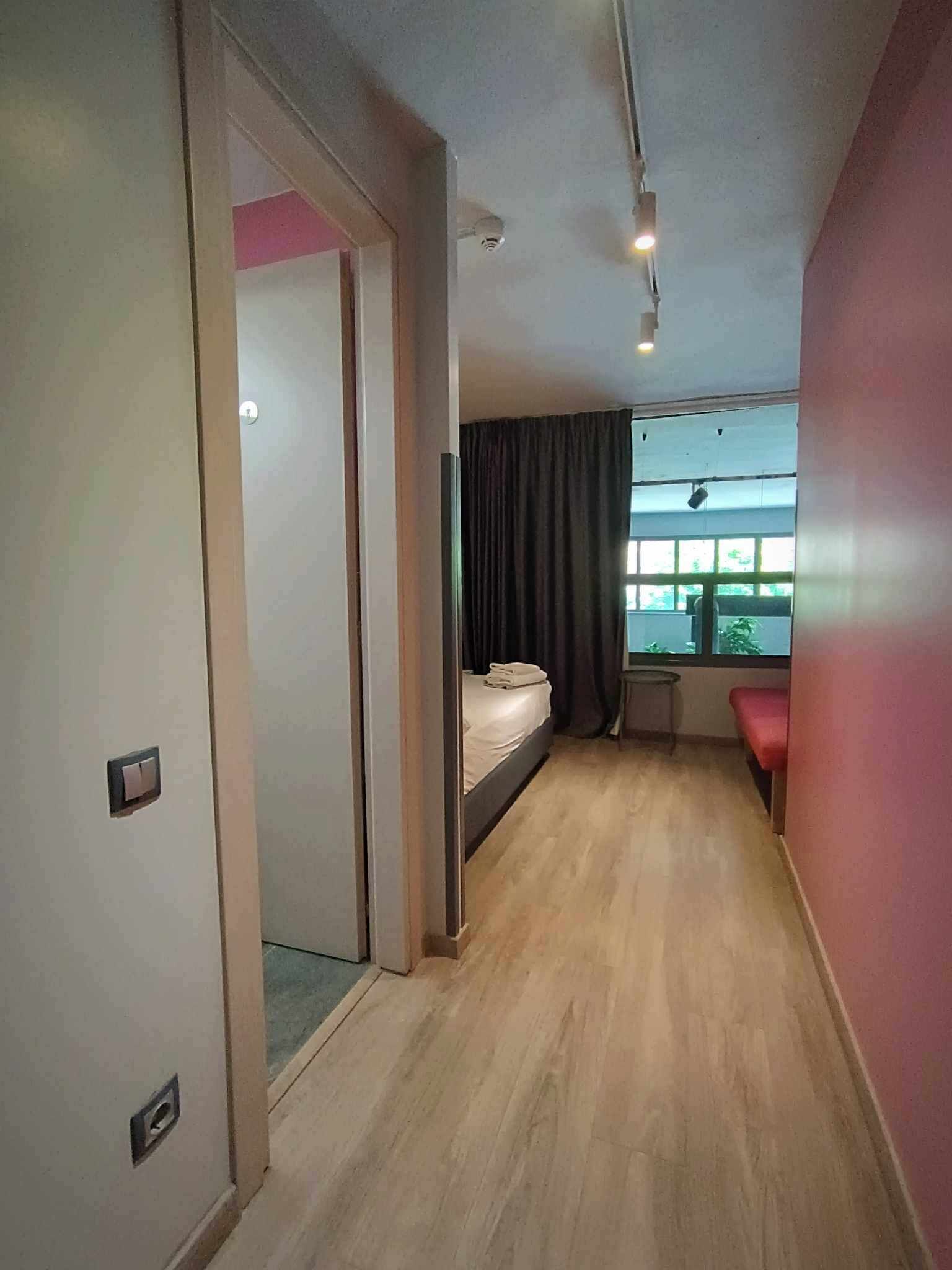A hotel room with a bed, a small table, a window with curtains, and a door leading to another room.
