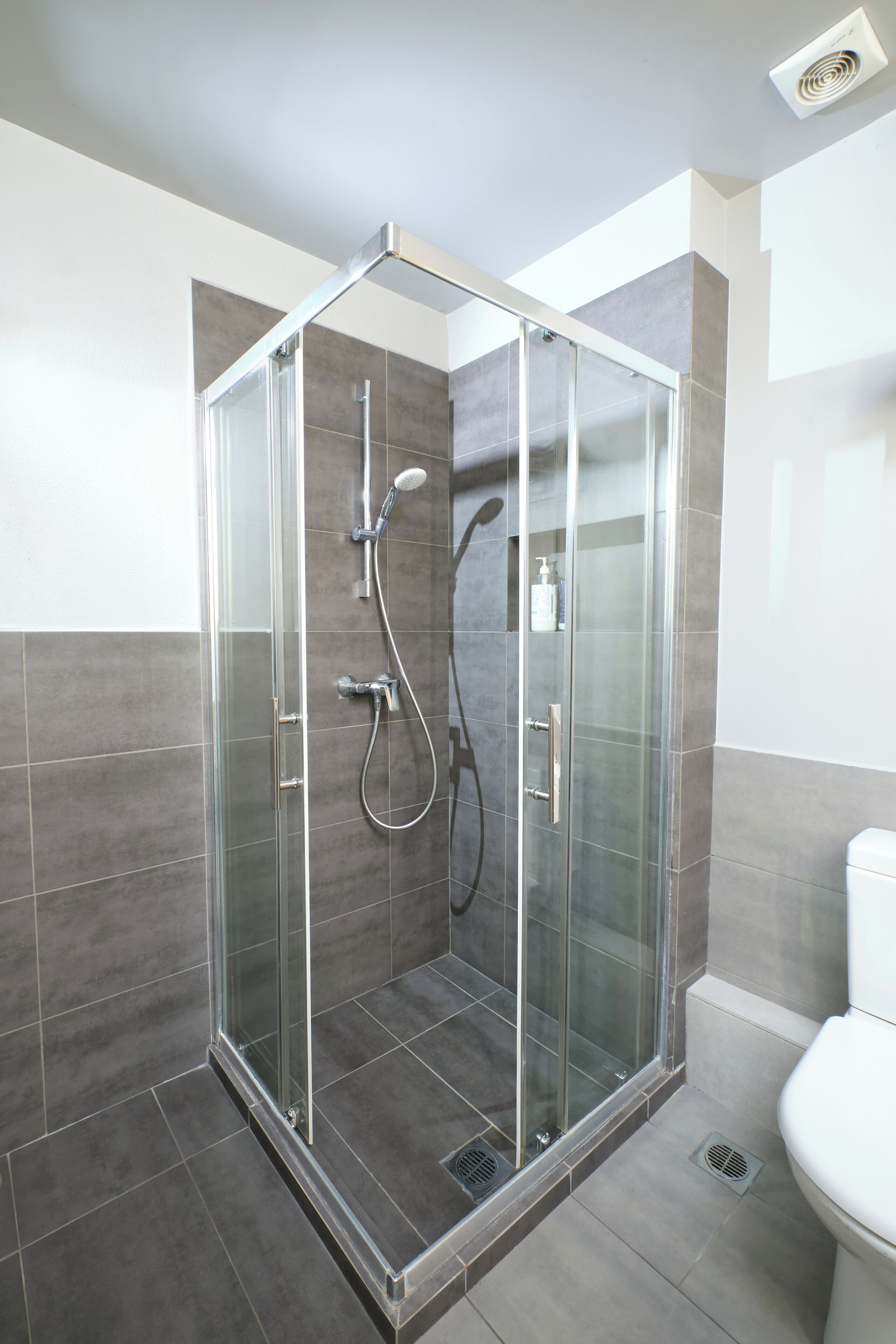 A modern bathroom with a glass-enclosed shower, grey tiles, and a white toilet.