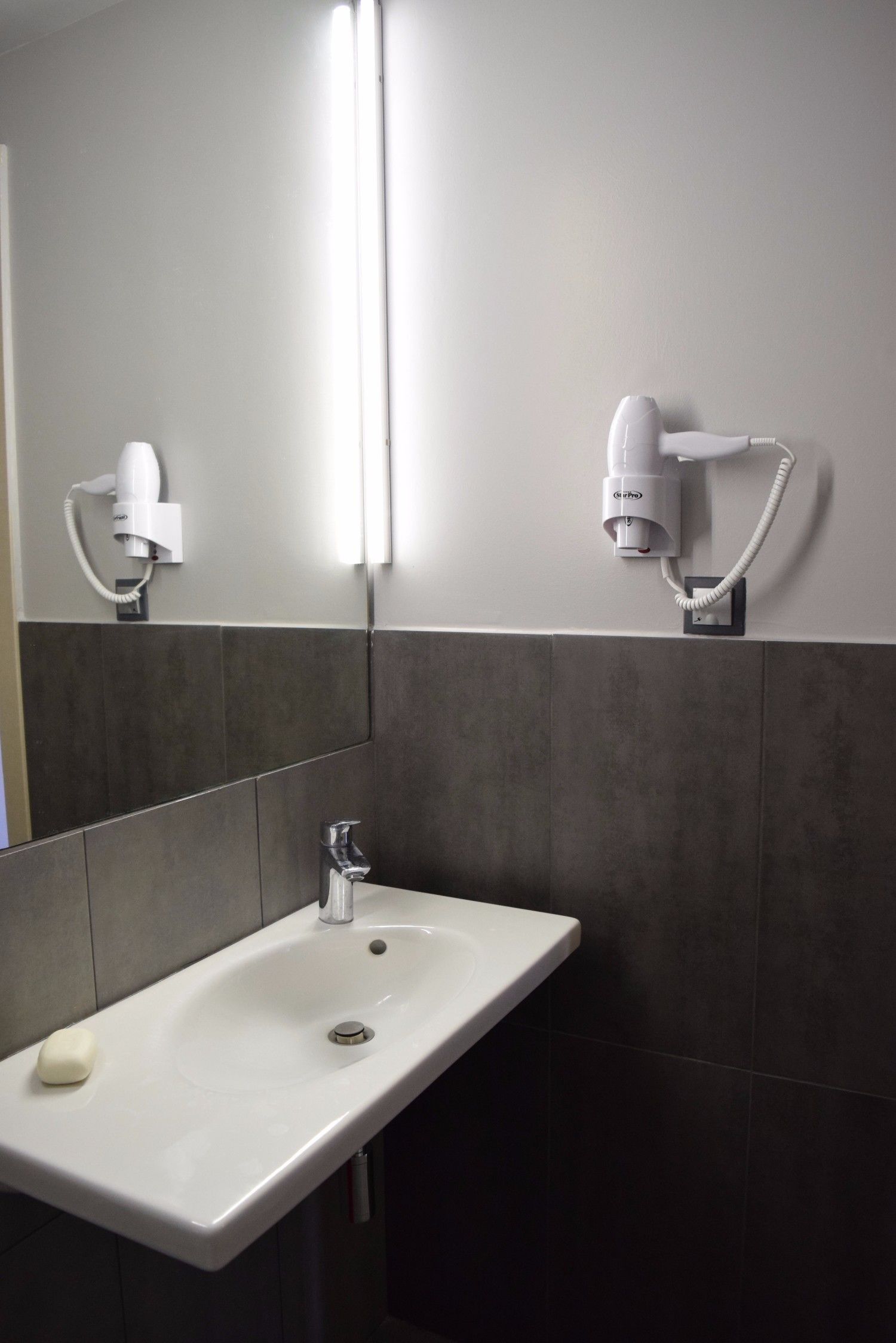 Une salle de bain avec un lavabo blanc, un miroir et deux sèche-cheveux fixés au mur.