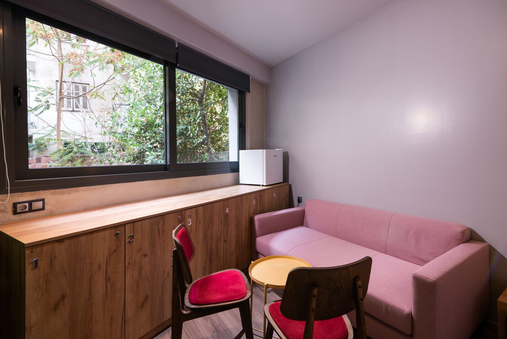 A compact room with a wooden desk, a pink sofa, and two chairs. A small table is placed between the sofa and the desk. A white appliance is visible on the desk.