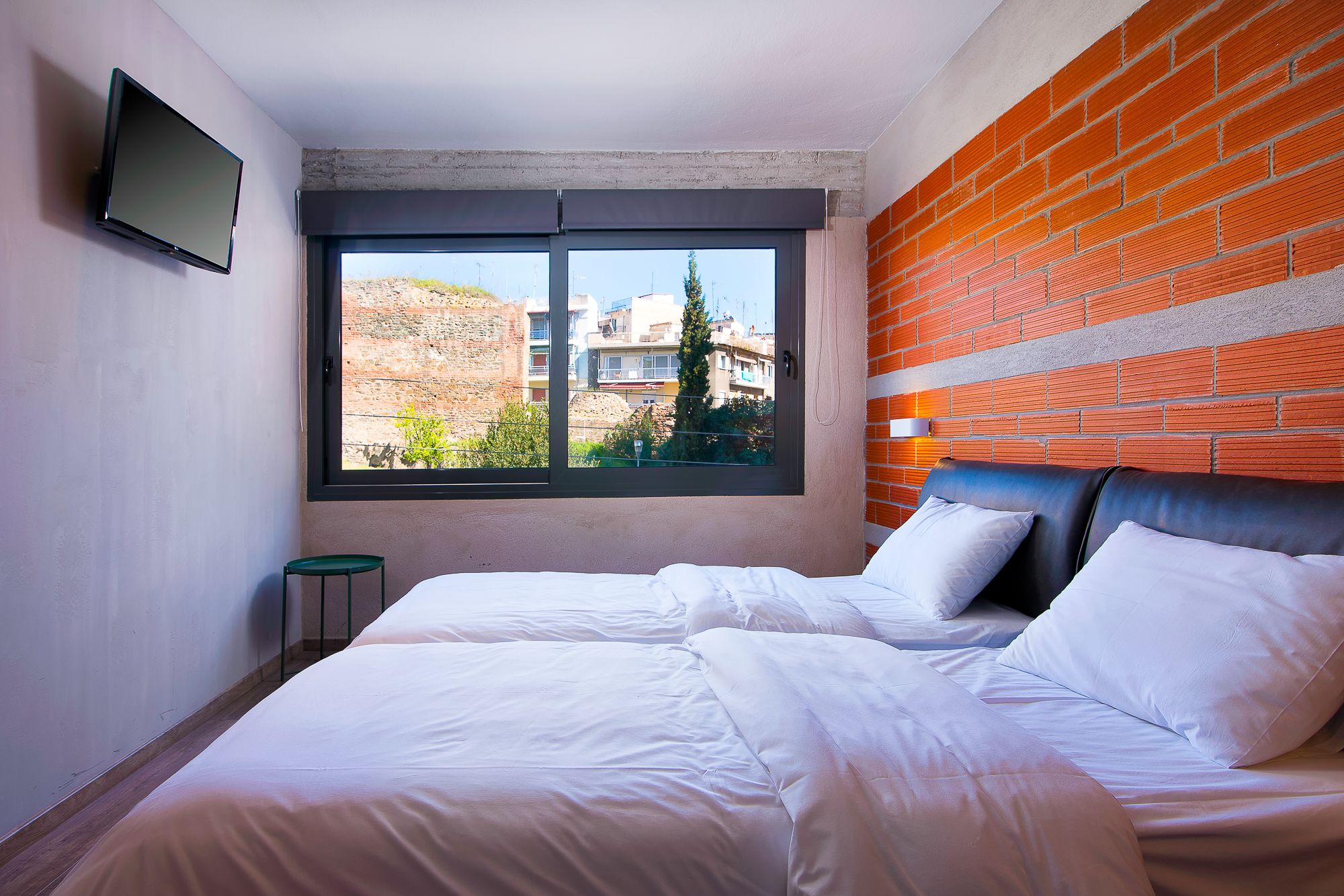 A modern hotel room with a large window, a brick accent wall, and a bed with white linens.