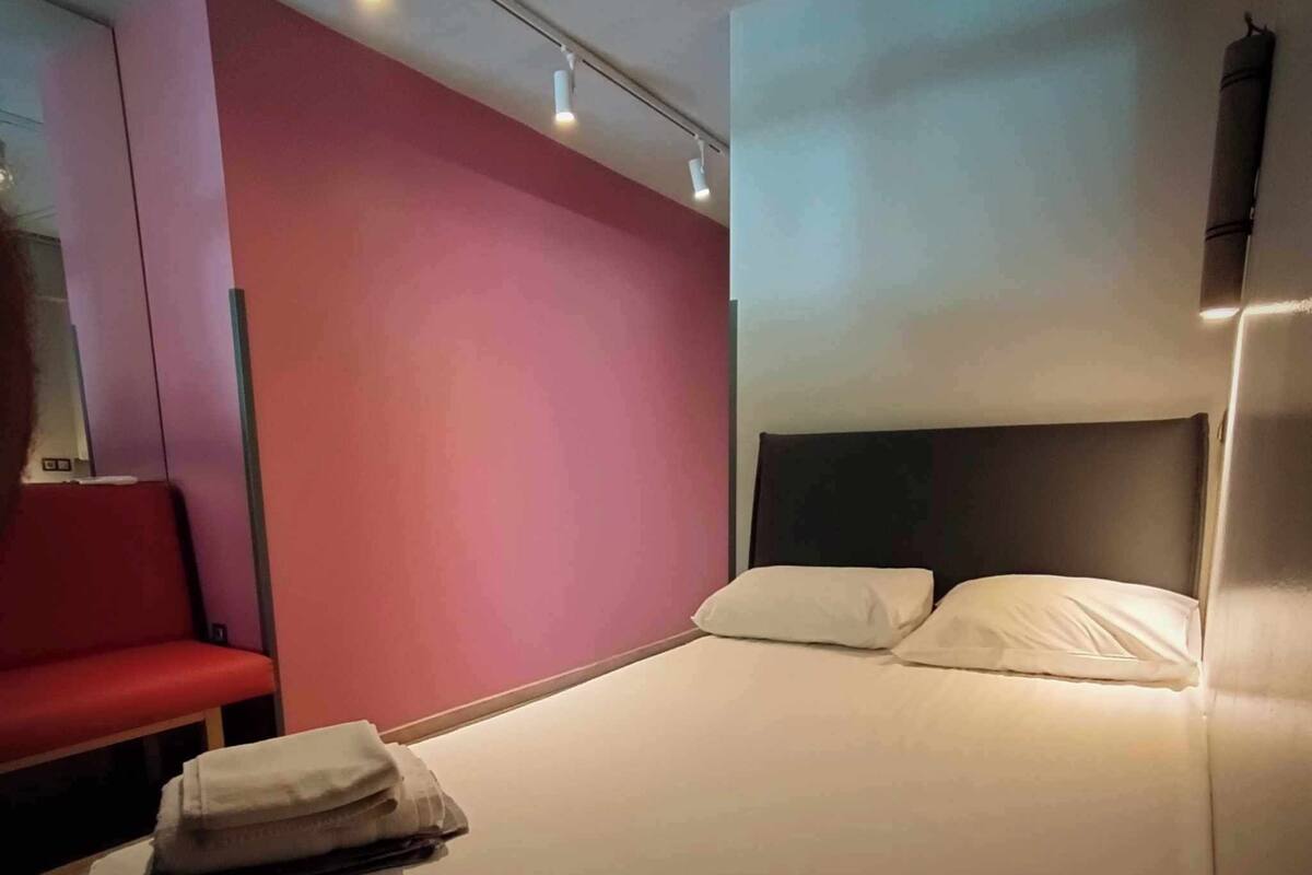 A hotel room with a bed, a red chair, and a wall-mounted light fixture.