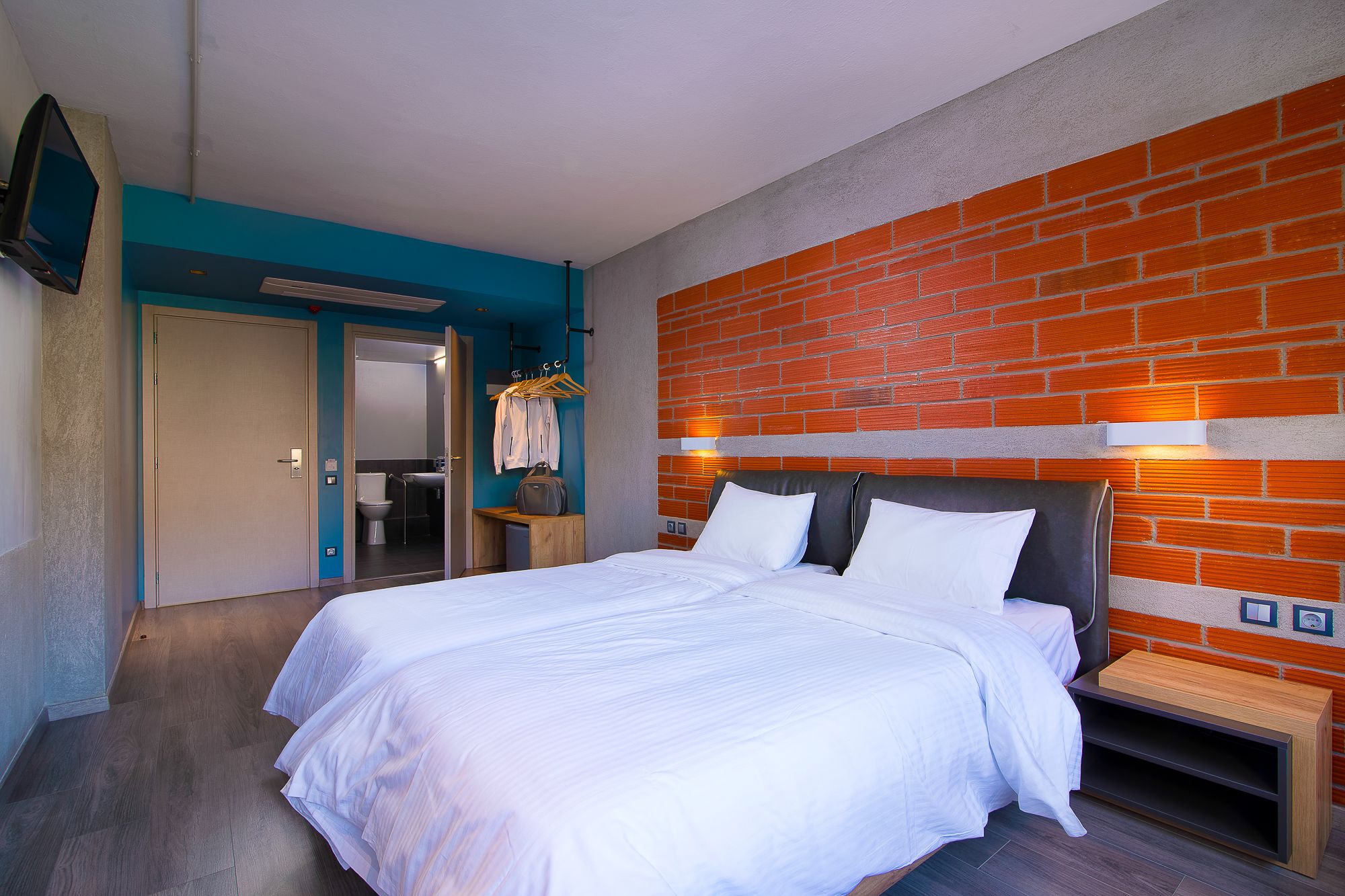 Habitación de hotel con una cama grande, una mesita de noche de madera, un muro de ladrillo decorativo, un baño visible a través de una puerta entreabierta y un perchero con ganchos.