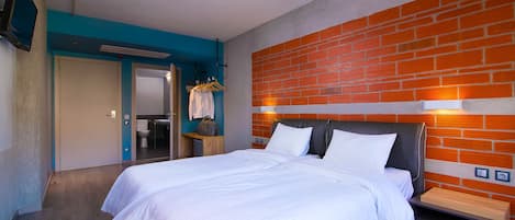 Une chambre d’hôtel avec un grand lit, une table de chevet en bois, un mur décoratif en briques, une salle de bain visible à travers une porte ouverte et un porte-manteau avec des cintres.