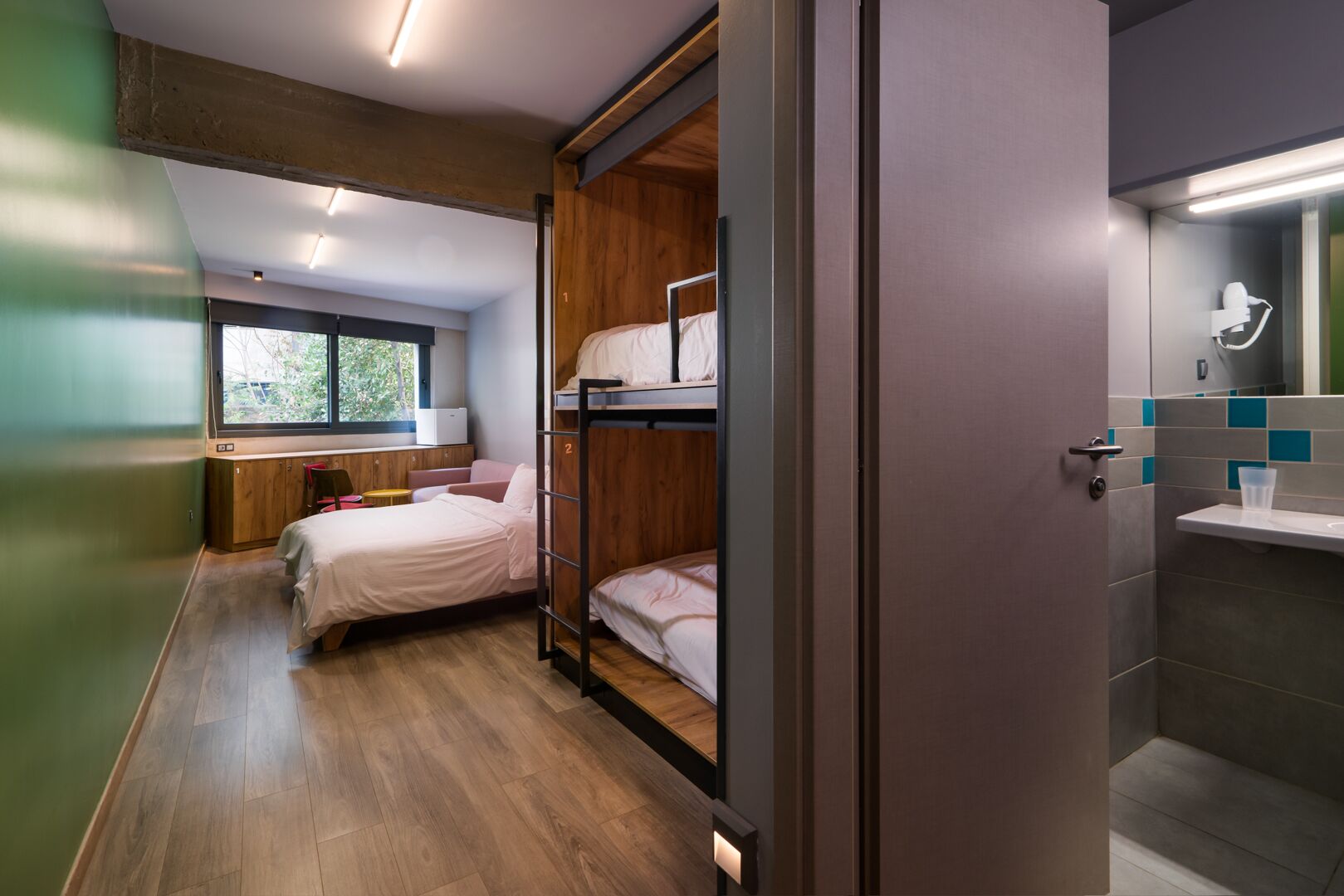 Una habitación de hotel moderna con literas, piso de madera, ventana con vistas y baño con espejo y lavamanos.