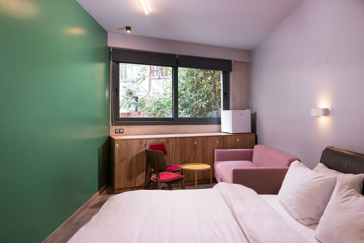 A hotel room with a bed, a seating area with chairs, a small table, a window with a view of greenery, and a wall-mounted light fixture.