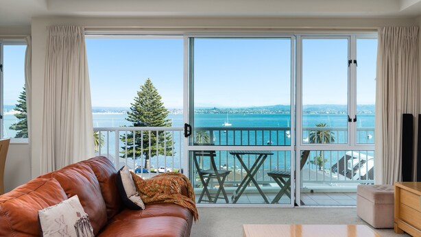 A modern living room with a leather sofa, a wooden coffee table, and a balcony with a view of the sea and city skyline. - The Beaumont Apartments (Tauranga)