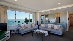 A living room with a sofa set, a coffee table, and a TV. There is a kitchen area with cabinets and a refrigerator. - The Beaumont Apartments (Tauranga)