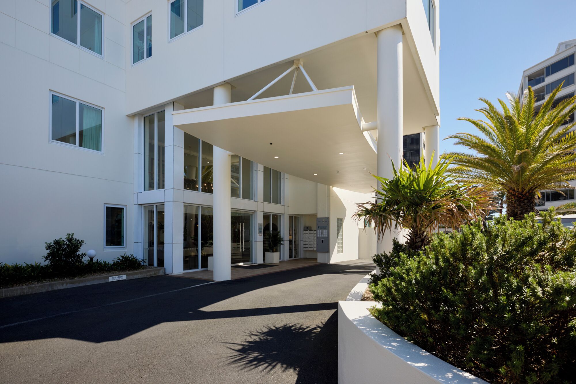 A modern building with a covered entrance, large windows, and a landscaped area with palm trees.