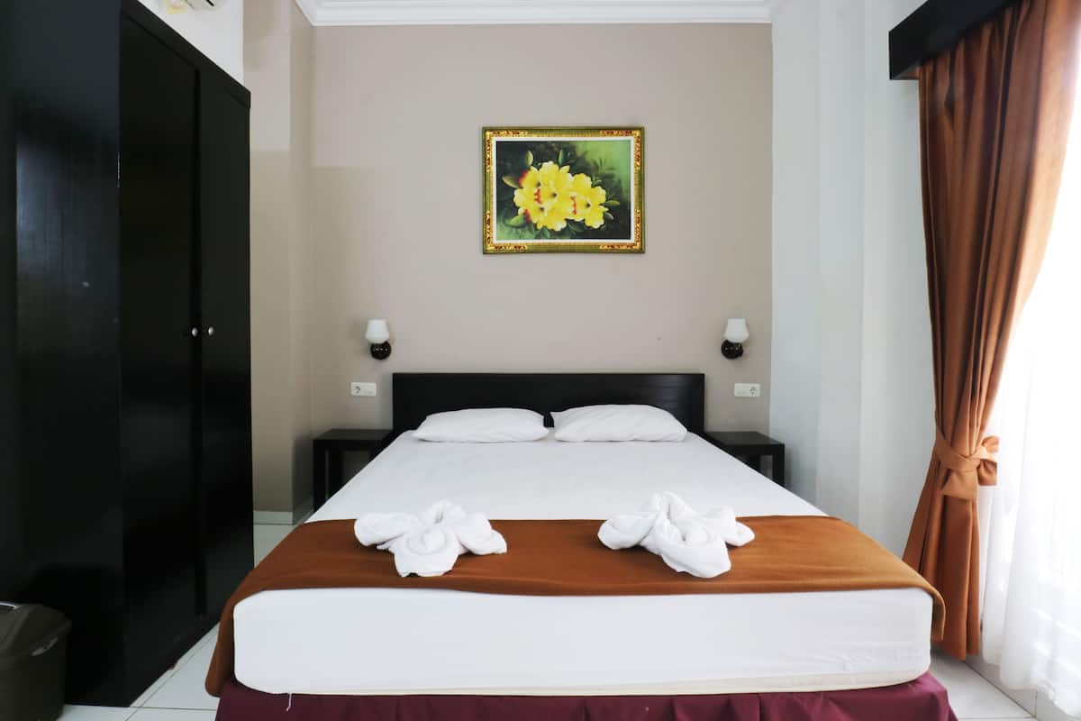 A hotel room with a large bed, two pillows, a brown headboard, a painting on the wall, a nightstand, a lamp, and a curtain.