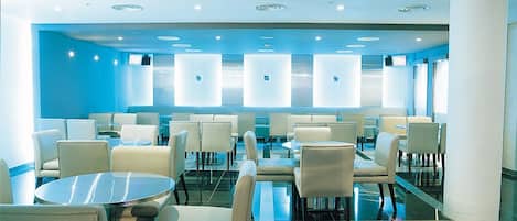 A modern hotel lobby with round tables and chairs, a glass partition wall, and a blue accent wall.