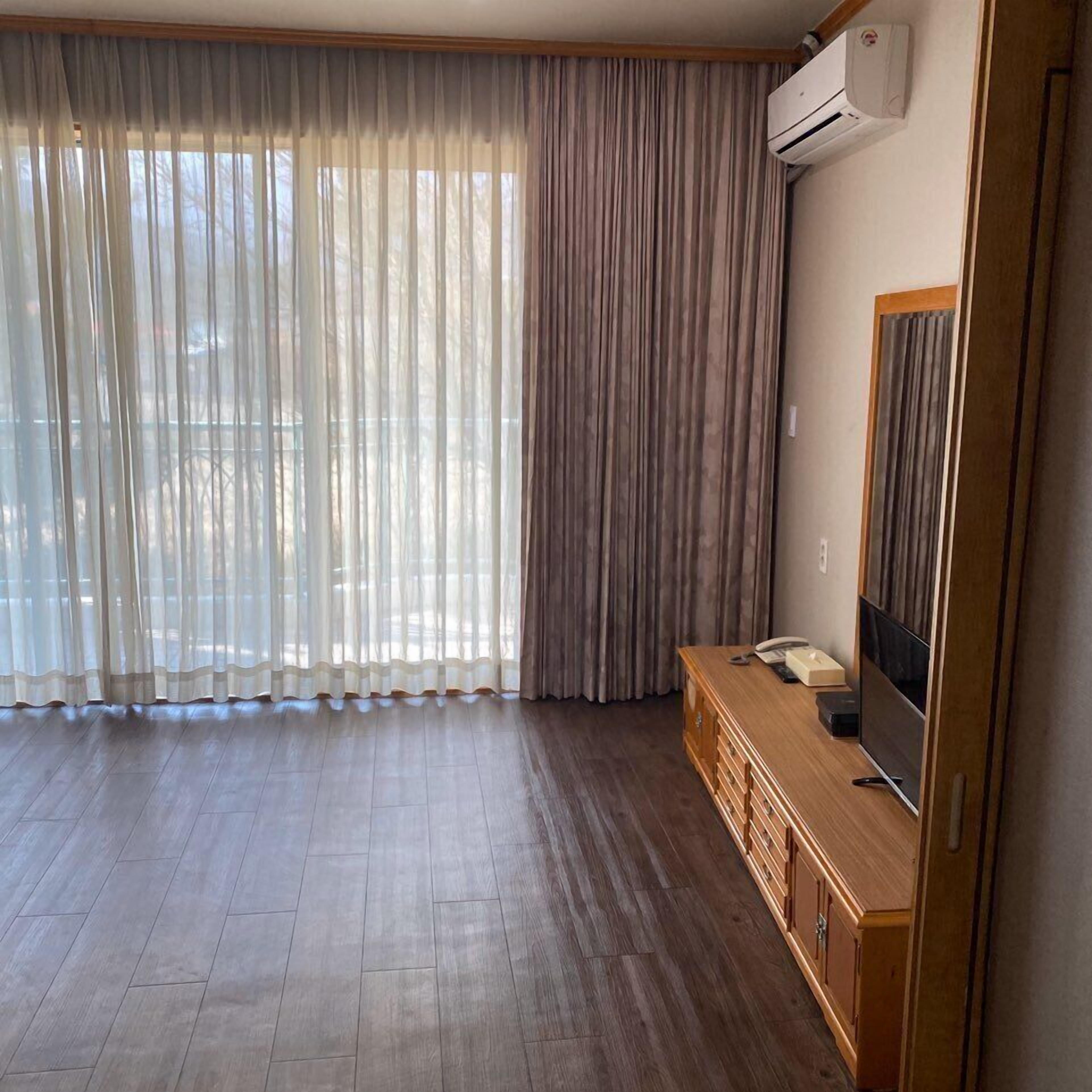 A room with a wooden floor, a TV on a wooden cabinet, and a large window with sheer curtains.