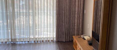 A room with a wooden floor, a TV on a wooden cabinet, and a large window with sheer curtains.