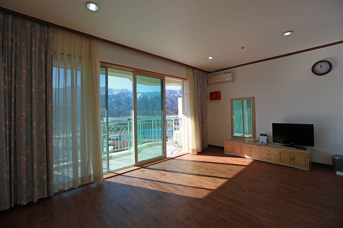 A spacious living room with a large sliding glass door leading to a balcony, a flat-screen TV on a wooden cabinet, and a wall-mounted clock.