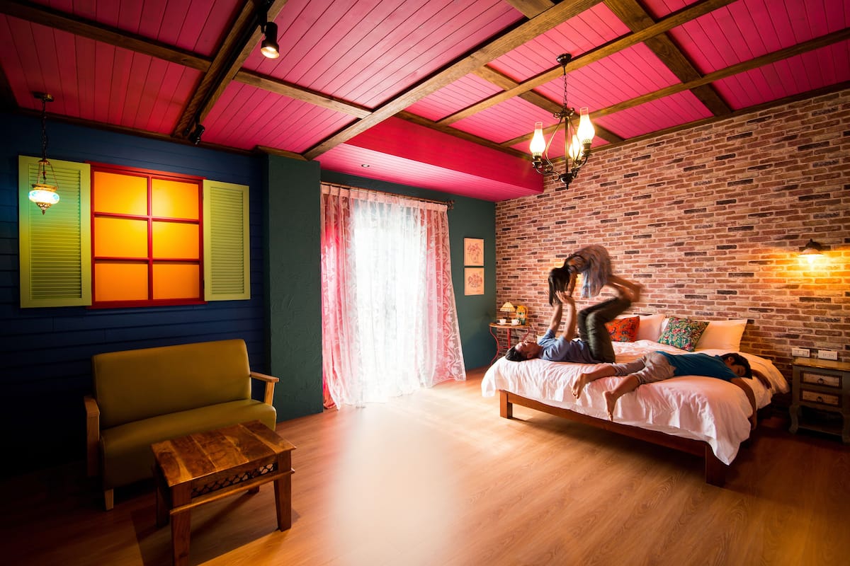 A room with a bed, a wooden bench, a window with shutters, and a brick wall.