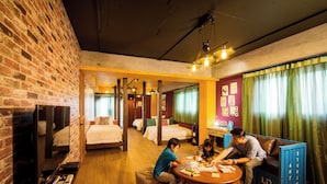 Habitación de hotel con pared de ladrillo, suelo de madera, una mesa redonda de madera y un sofá. Se ven camas al fondo y un televisor a la izquierda.