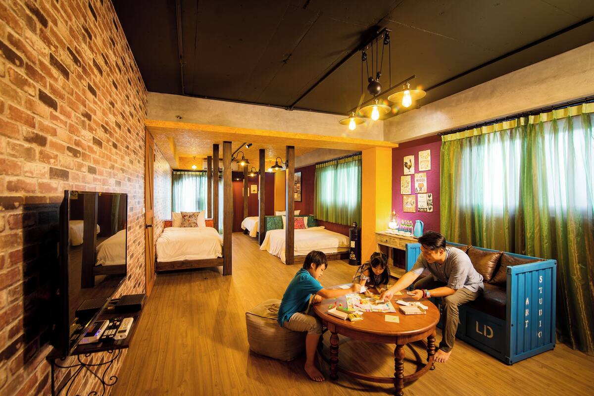 A hotel room with a brick wall, wooden floors, a round wooden table, and a sofa. There are beds in the background and a television on the left.