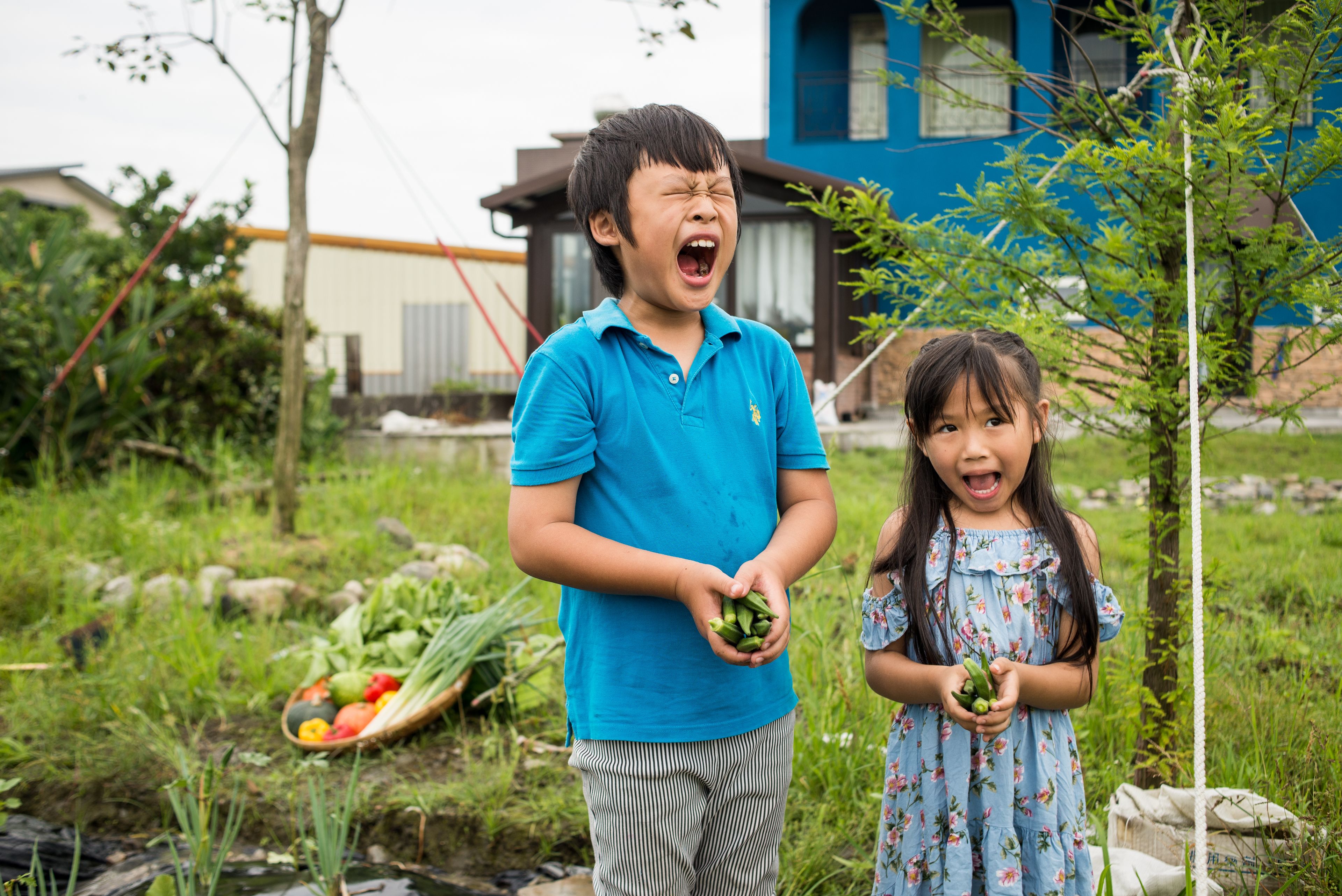 Un niño y una niña sostienen verduras en un jardín, con una casa al fondo.
