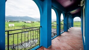 Un balcón techado con columnas azules y vista a un paisaje verde y montañas.