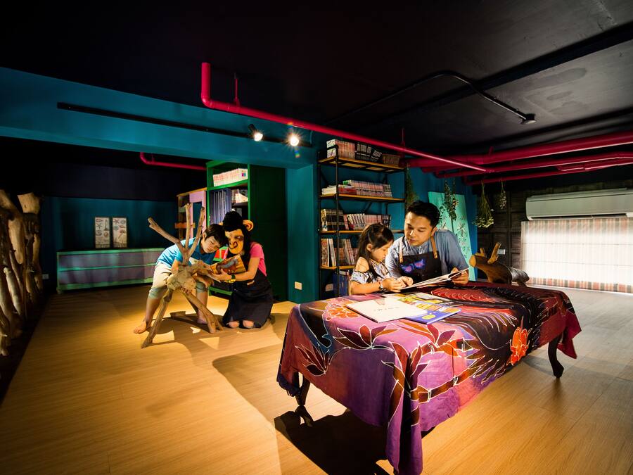 A room with a wooden floor, a table with a colorful tablecloth, and a bookshelf filled with books.