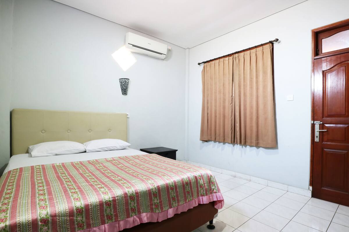 A bedroom with a bed, a nightstand, an air conditioning unit, a window with curtains, and a wooden door.