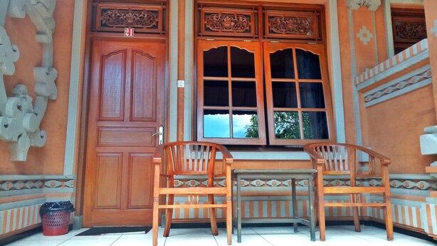 A traditional wooden door with intricate carvings, a window with a grid pattern, and a small table with two chairs in front. - Taman Mekar Beach Inn (Kuta)