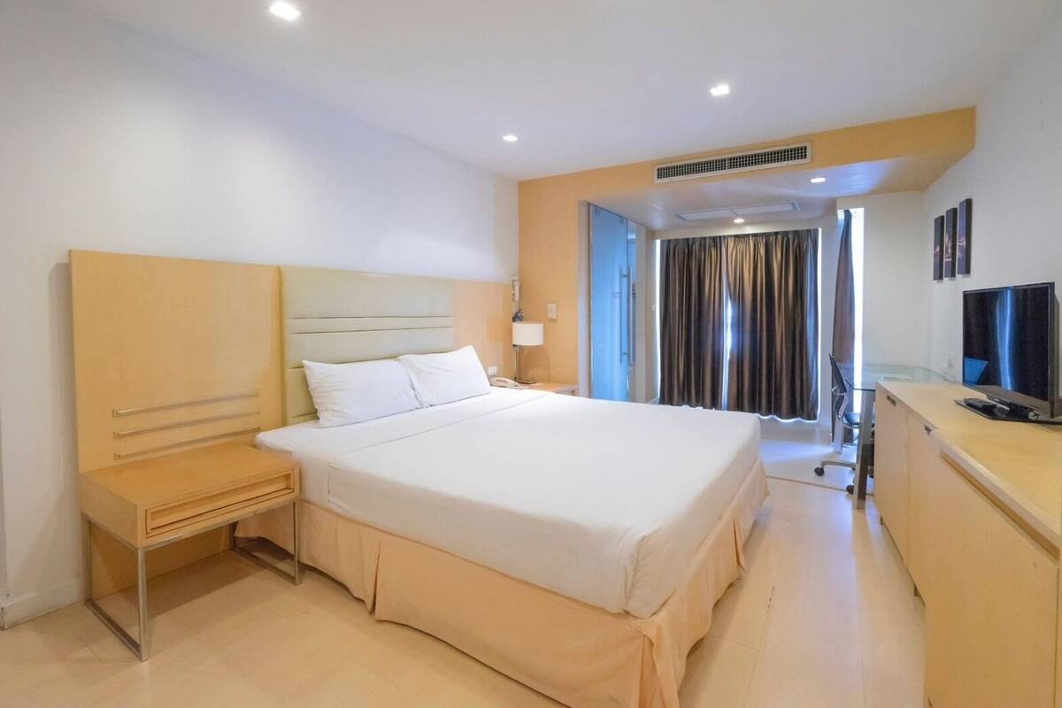 Une chambre d’hôtel comprenant un lit, une table de chevet, un bureau, une télévision et une fenêtre avec des rideaux.