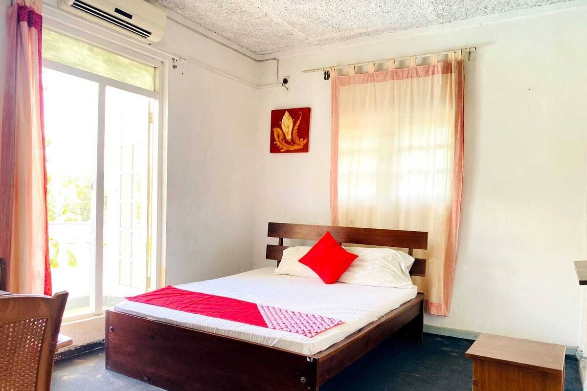 Una cama individual con cabecera de madera, almohada roja y colcha blanca en una habitación con ventana, cortina e imagen colgada en la pared.