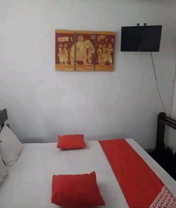 A simple room with a bed, pillows, a TV mounted on the wall, and a painting above the bed.