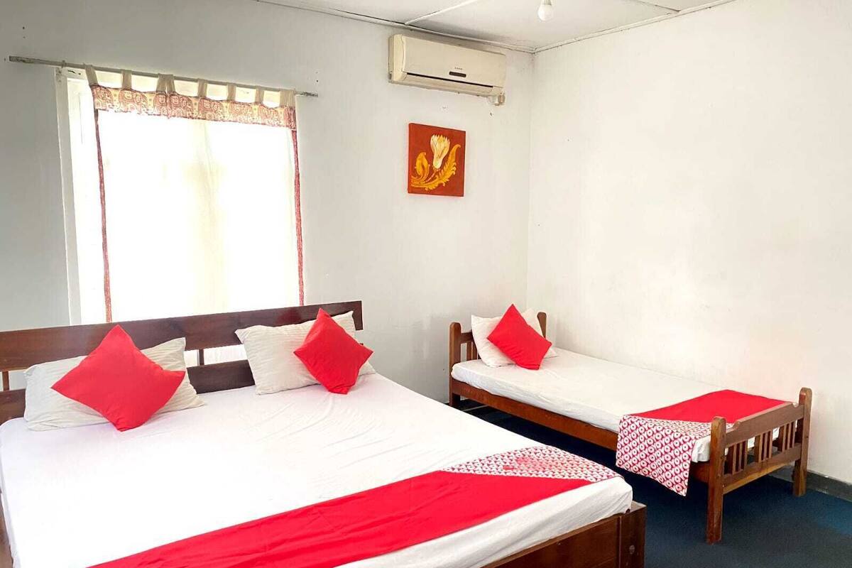 Una habitación con dos camas individuales, cada una con una almohada roja y una colcha blanca. Hay una ventana con cortina, un equipo de aire acondicionado en la pared y un pequeño cuadro rojo y amarillo sobre las camas.