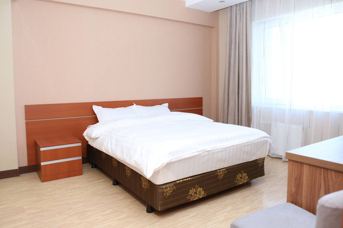 Una cama bien hecha con cabecera de madera, una mesita de noche con cajones y una ventana con cortinas.