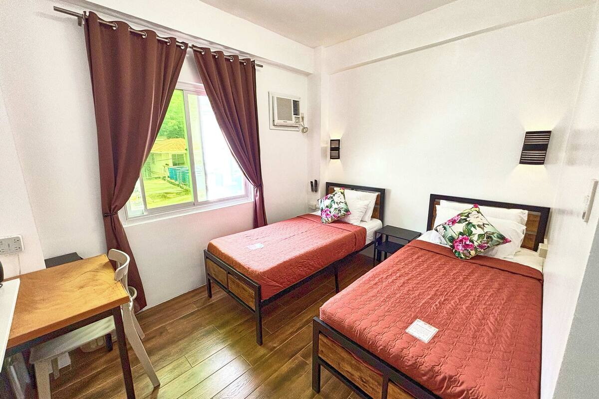 A room with two single beds, a wooden table, a chair, a window with curtains, and a small table with a lamp.
