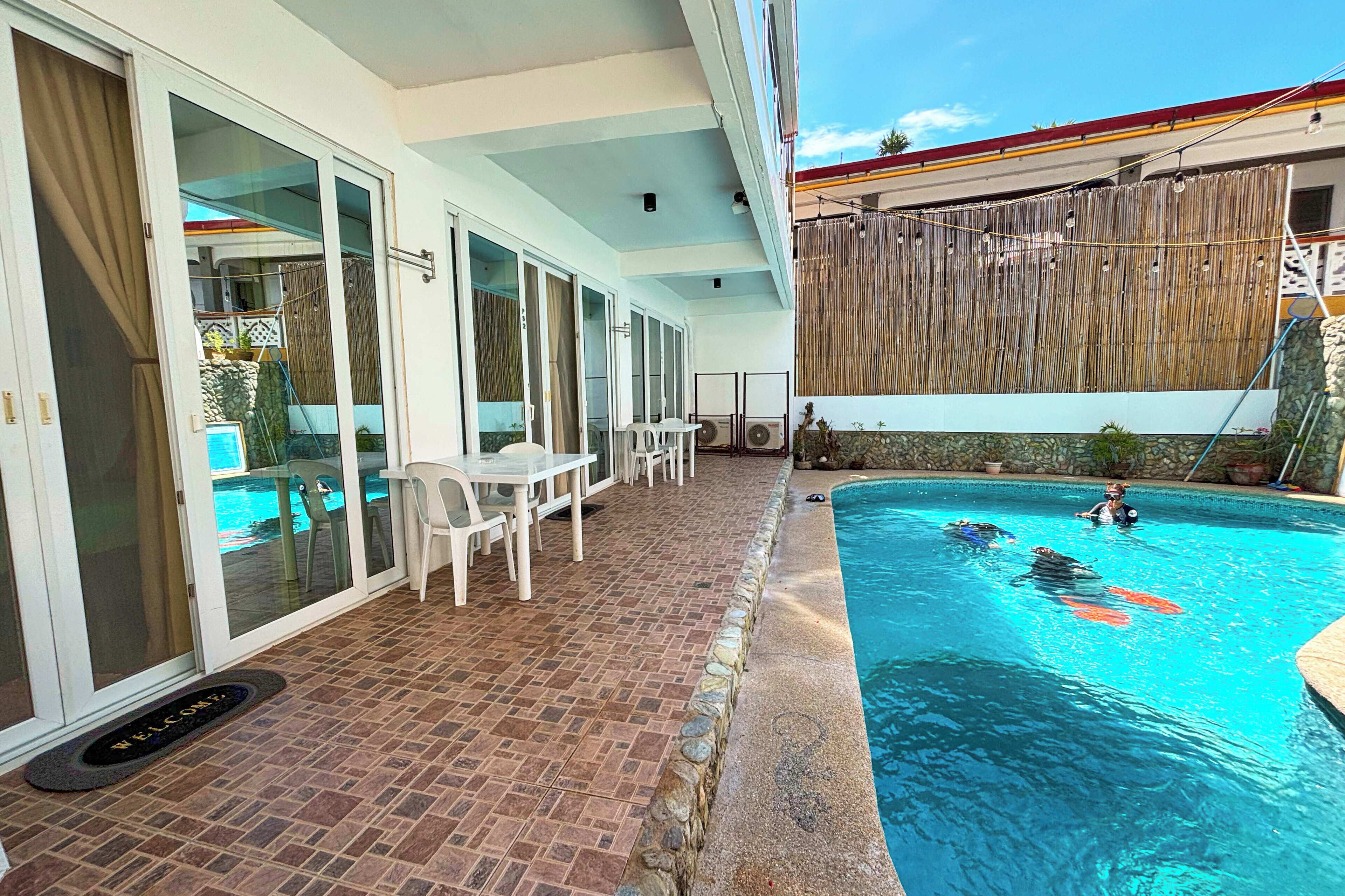 A swimming pool with a tiled deck, a covered patio area with a table and chairs, and a wooden fence in the background.